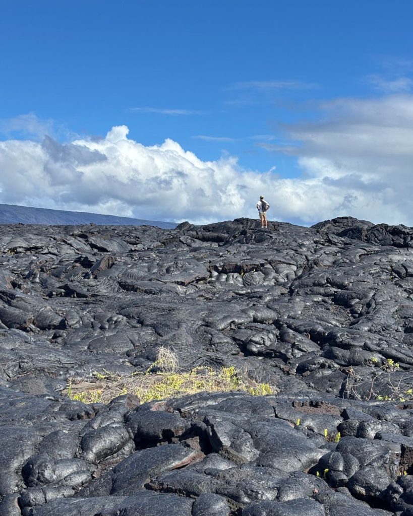 A lone hiker standing on a vast field of black pahoehoe lava under a blue sky