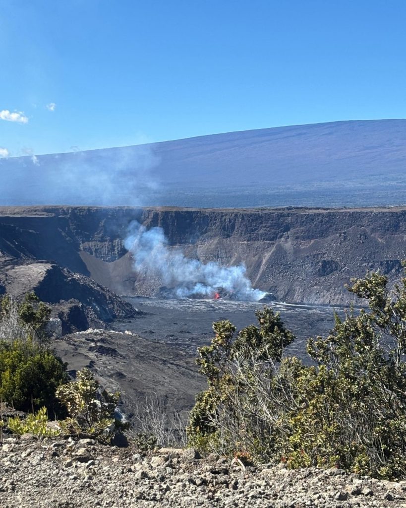 Steam and a patch of glowing lava inside Kilauea crater, seen from the rim with Mauna Loa in the distance