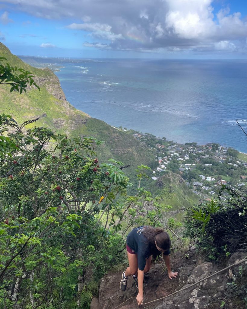 Hiker scrambling up a rope on a steep ridge on the Kuliouou Ridge Trail with an ocean view below