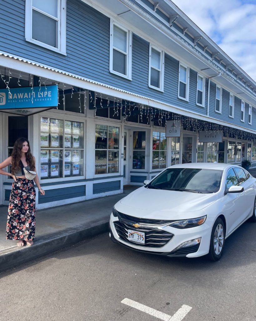 A white rental car parked outside a blue two-story wooden storefront building in a small Big Island town