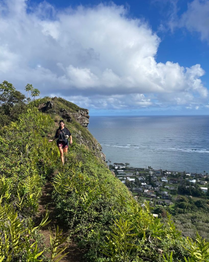 Hiker walking along a narrow grassy ridge line on the Kuliouou Ridge Trail with the ocean and clouds in the distance