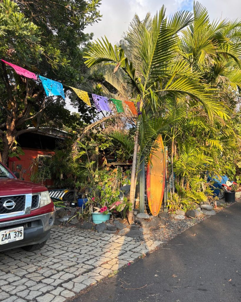 An orange surfboard and a string of colorful flags in a palm-filled front yard next to a parked pickup truck big island itinerary