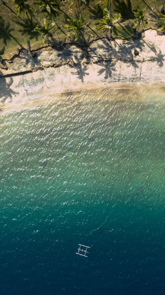 Aerial view of a single outrigger boat on turquoise water off the tree-lined shore of Gili Asahan