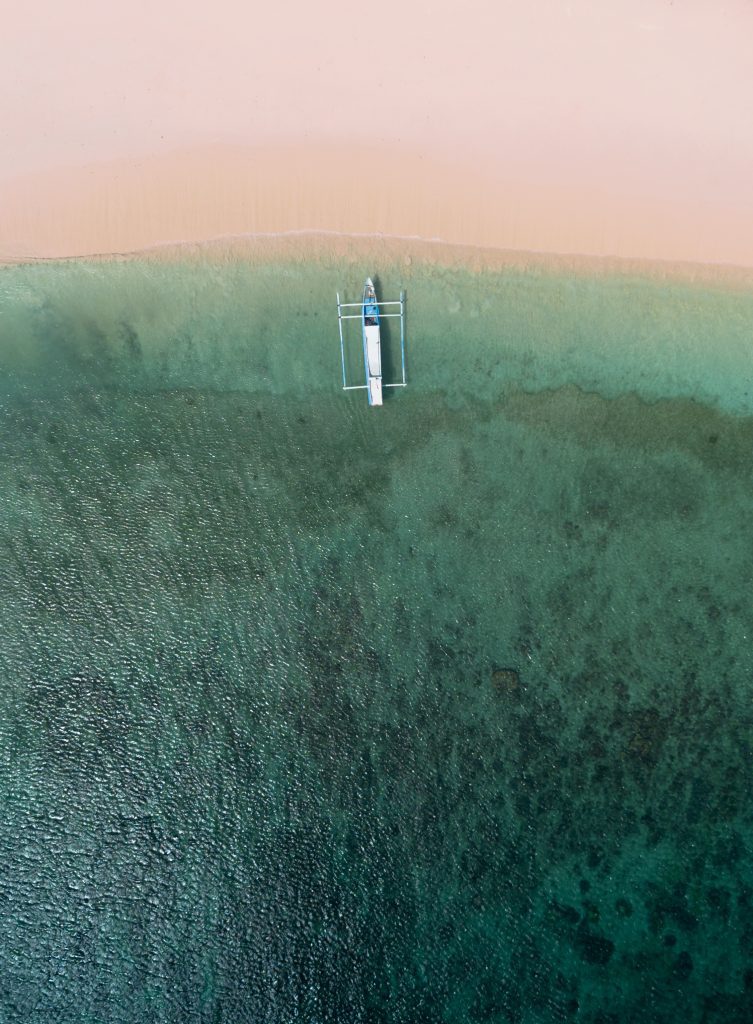Aerial view of Pink Beach, with pink sand, a dark reef and one outrigger boat in the shallows