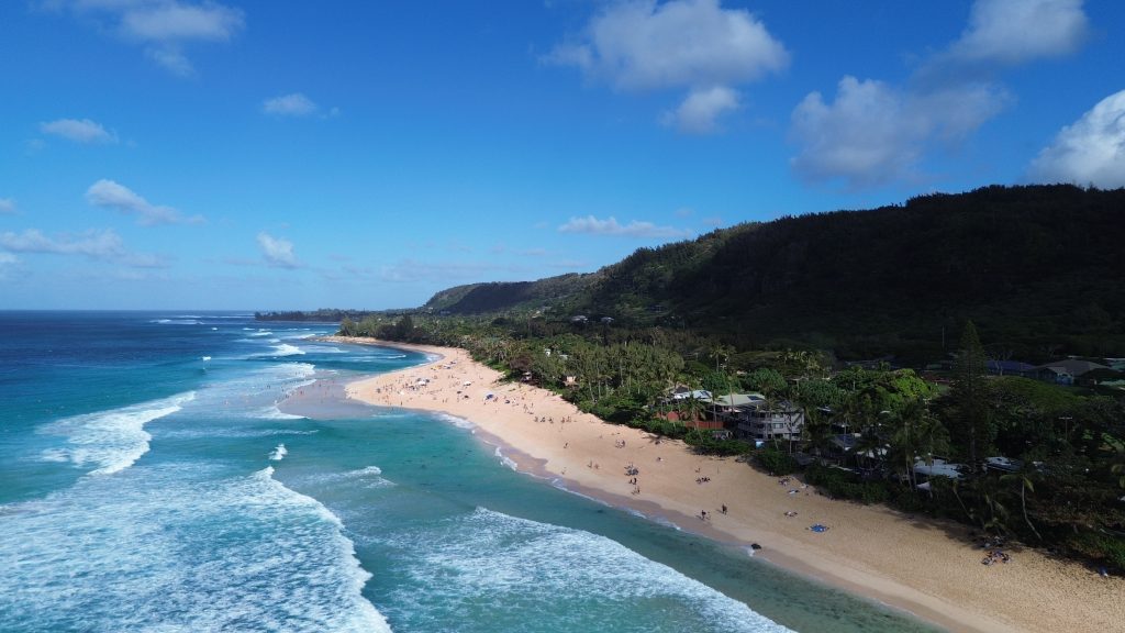 Aerial drone view of Oahu's North Shore coastline with turquoise water, breaking waves and a sandy beach lined with greenery