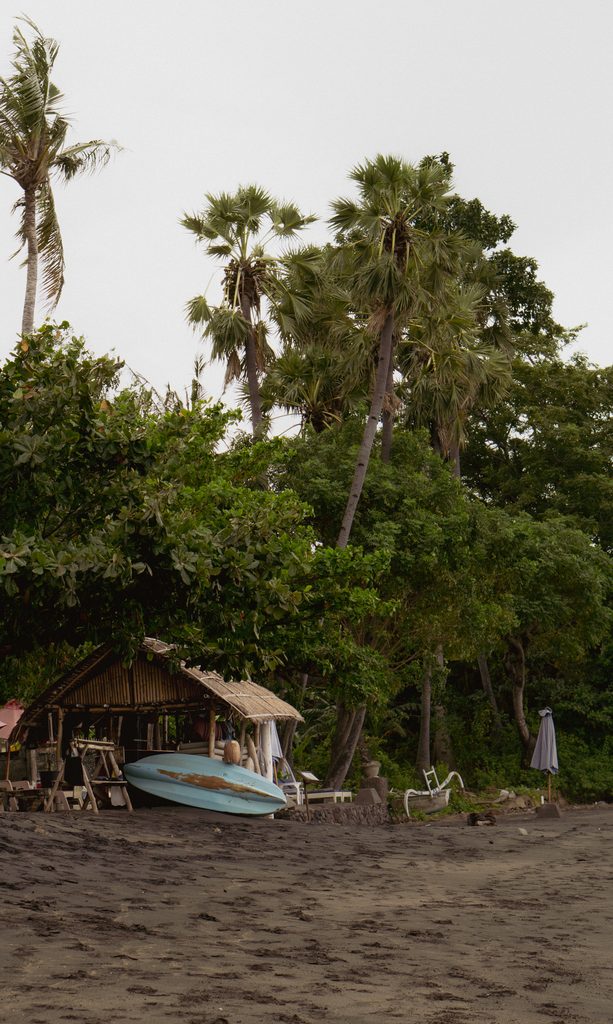 Wooden beach shack and a blue outrigger canoe under palm trees on the black sand at Pura Sentana Hill Beach, Amed