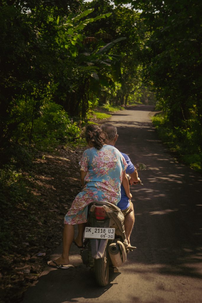 Two people riding a scooter along a shaded country lane through the trees near Amed