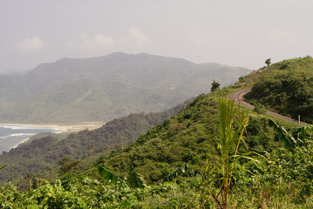 Coastal road curving along a green ridge above an empty white sand bay in Sekotong