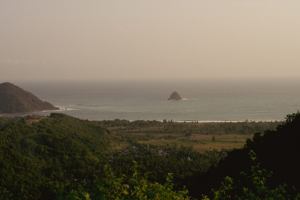 Hazy view over forested hills down to a wide bay in Lombok, with a small rocky island offshore