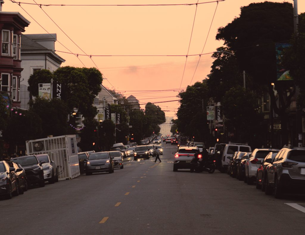 A steep San Francisco street lined with parked cars under an orange evening sky