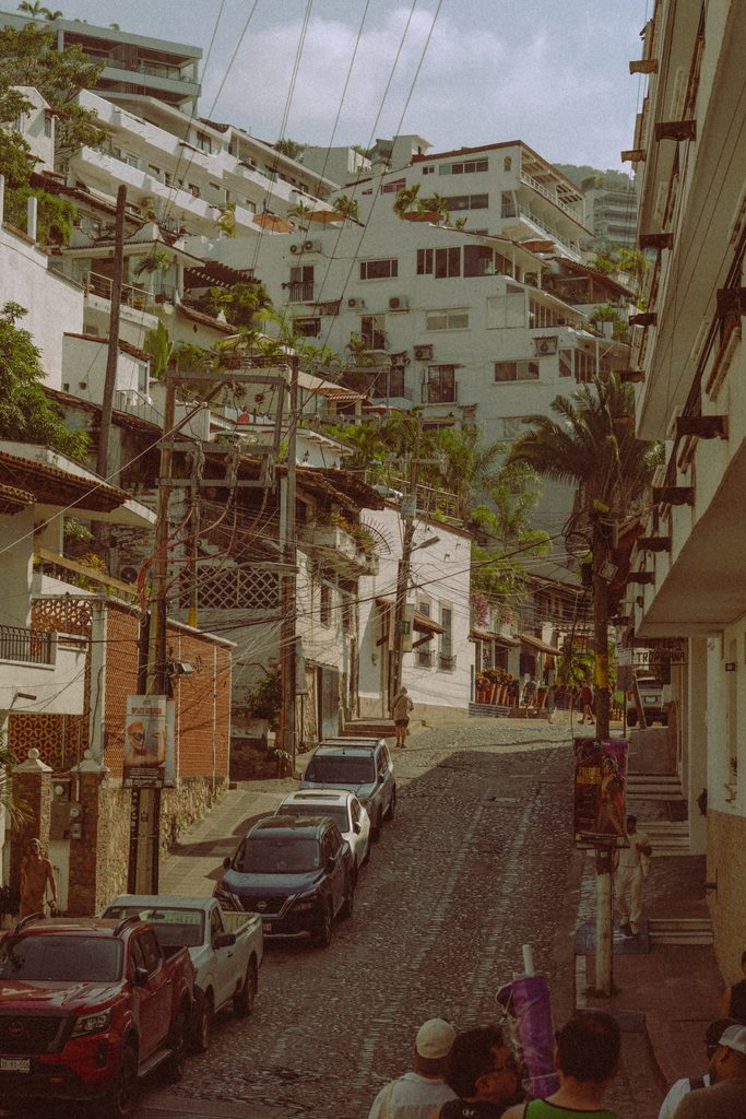 Steep residential street climbing the hillside in Puerto Vallarta, lined with parked cars, power lines and white apartment blocks