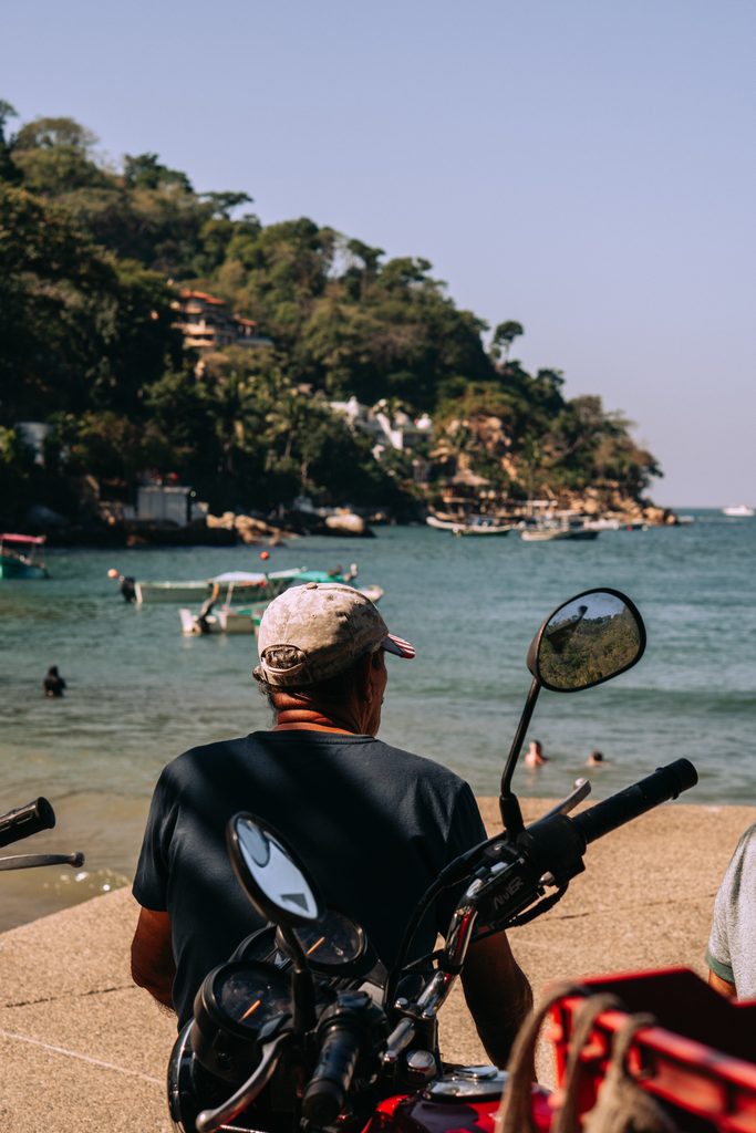 A man sitting on a motorbike on the beach at Boca de Tomatlan, looking out at the boats in the bay