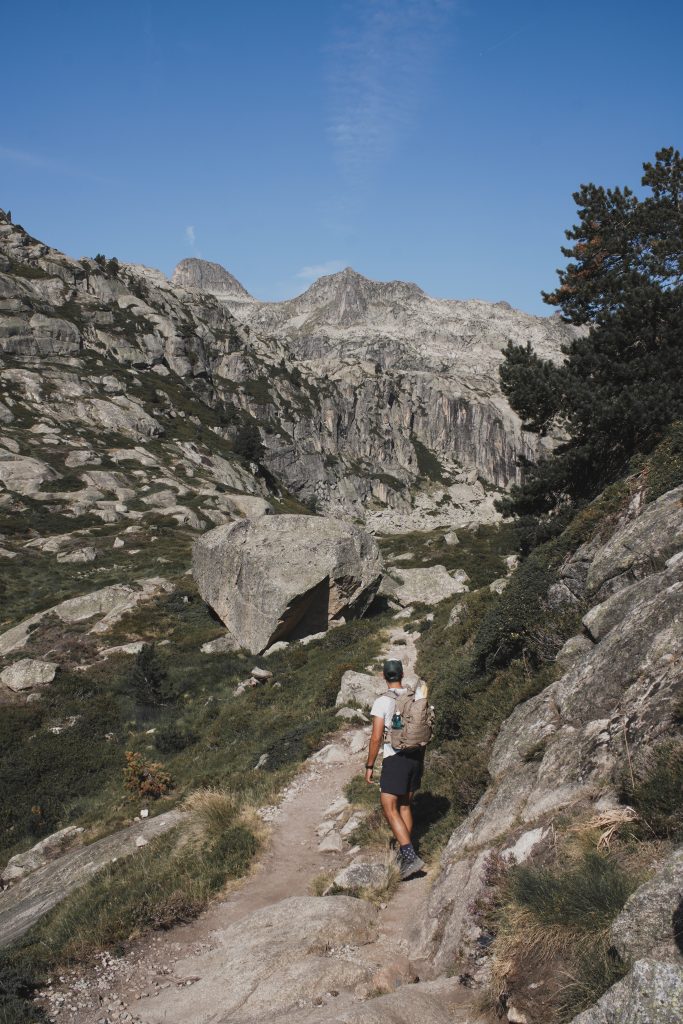 Randonneur marchant sur un sentier rocheux entre des pentes herbeuses, dans un vallon encaissé du Marcadau.
