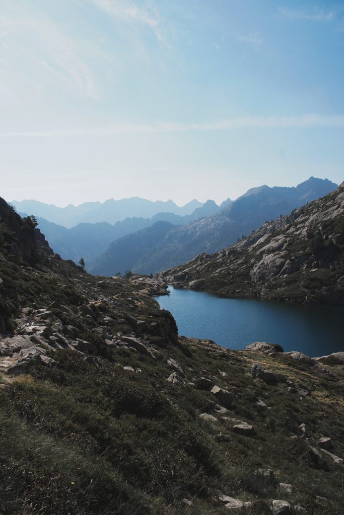 Vue plongeante sur un lac turquoise entouré de rives verdoyantes, avec une succession de crêtes pyrénéennes en arrière-plan.