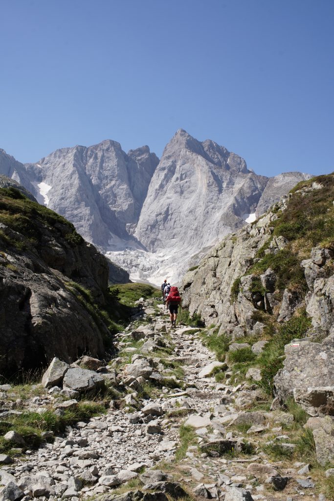 Hiker walking along a rocky mountain trail towards a large peak with patches of snow