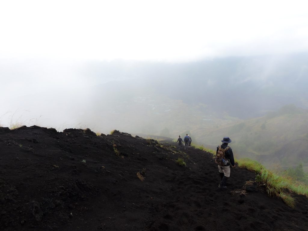 Hikers climbing the black volcanic slope of Mount Batur through mist at sunrise