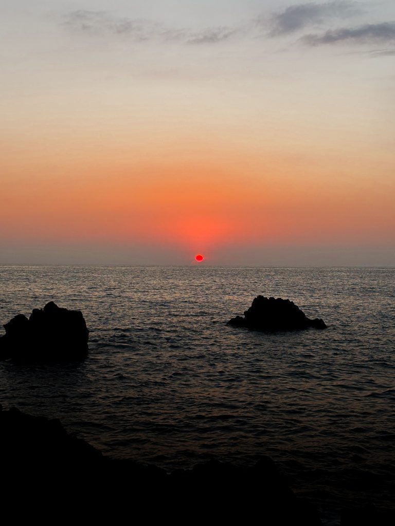 The sun setting into the ocean behind black lava rocks on the Kona coast