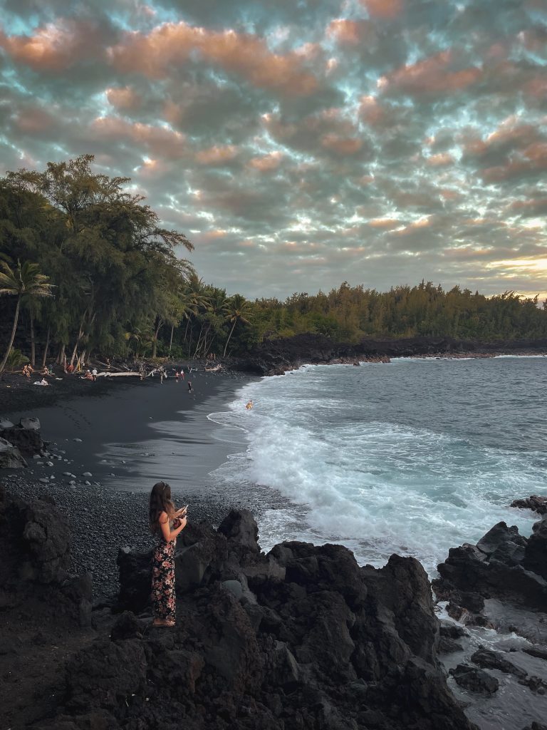 A person standing on lava rock above the black sand cove at Kehena Beach at dusk