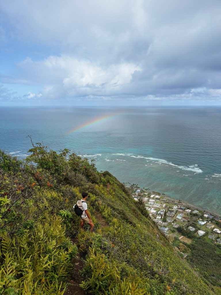 Hiker descending the Kuliouou Ridge Trail with a rainbow arching over the coastline and ocean below