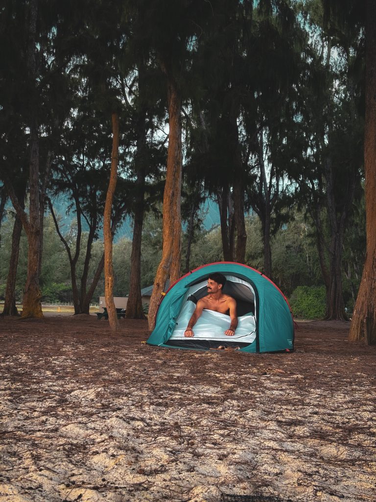 Man lying in a tent set up on sandy ground among tall ironwood trees at an Oahu campground