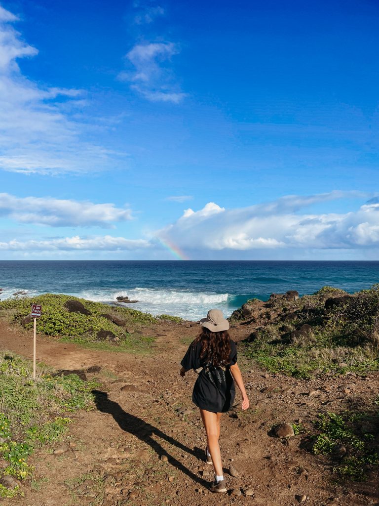 Woman walking a dirt coastal trail at Kaena Point with waves crashing on lava rocks and a faint rainbow in the sky