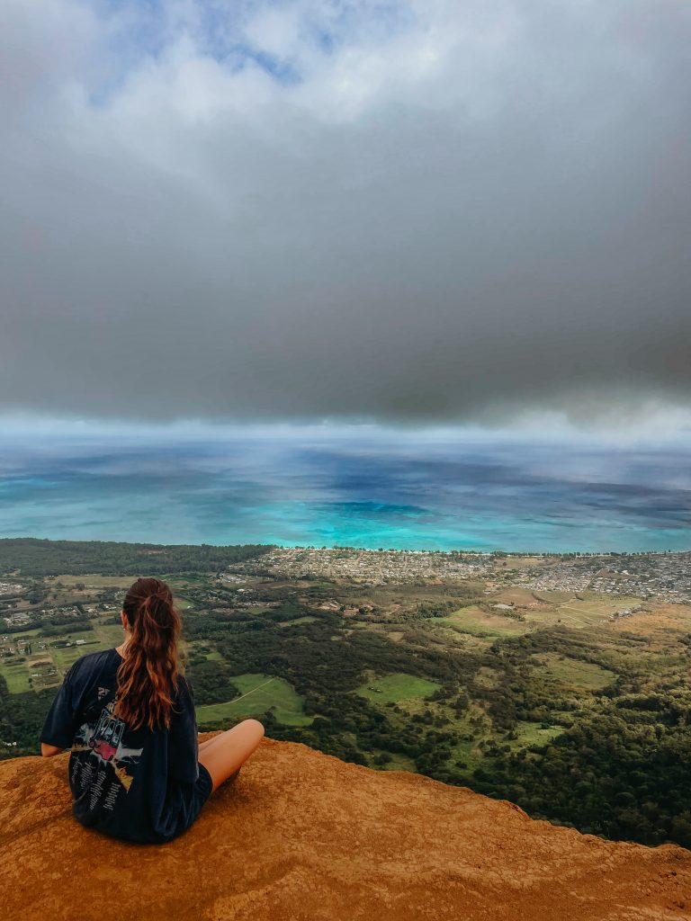 Woman sitting at the Ehukai Pillbox Trail lookout overlooking Lanikai and the east coast under a cloudy sky