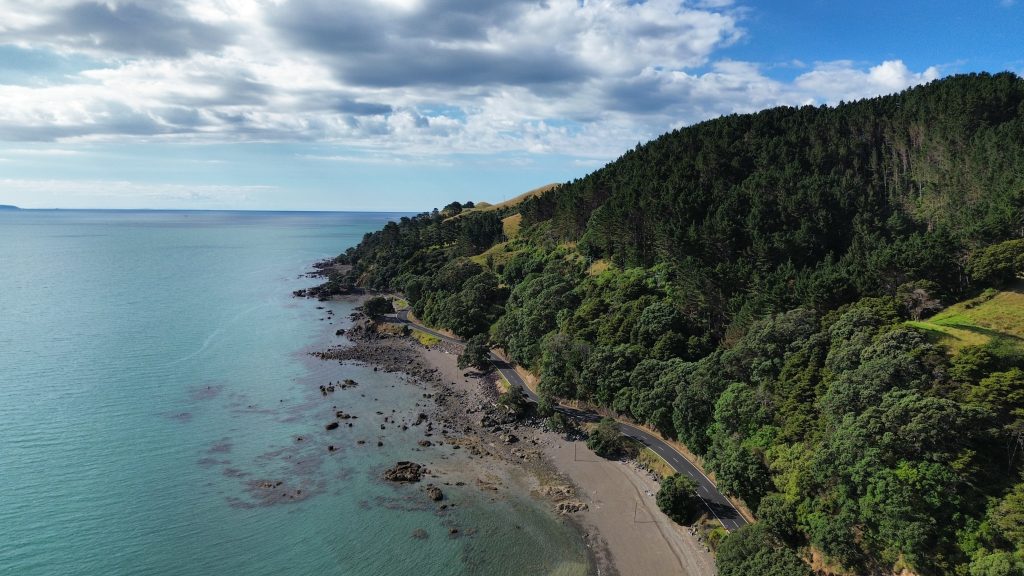 Aerial drone view of a coastal road winding along the Coromandel Peninsula coastline, with forested hills on one side and a rocky beach and turquoise sea on the other