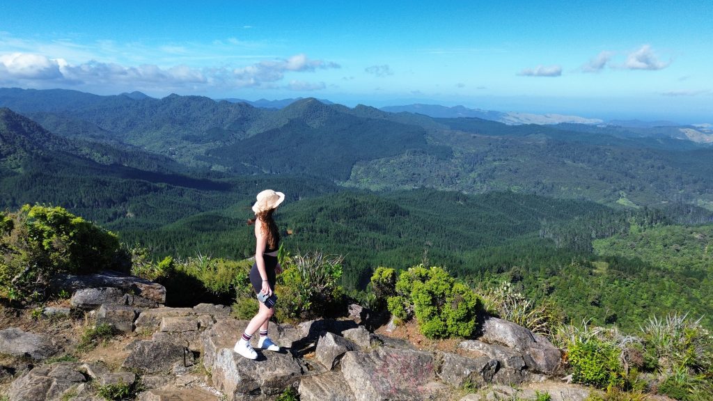 A woman standing on a rocky outcrop near Castle Rock, looking out over the forested hills of the Coromandel Peninsula