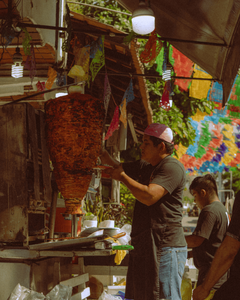 A taco stand with a vertical al pastor spit under strings of colourful papel picado bunting