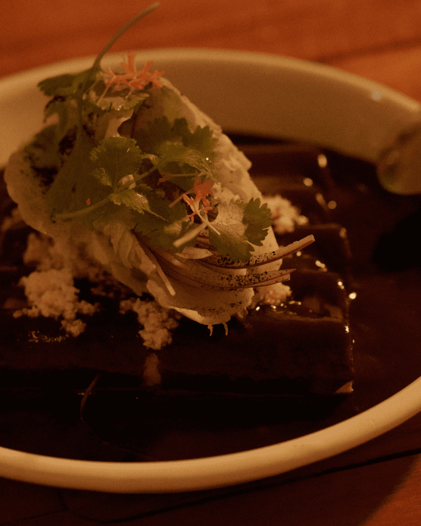 A dish covered in dark mole, topped with coriander and sliced red onion, in a dimly lit restaurant